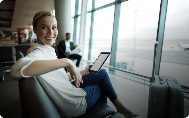 Woman sitting in an airport lounge with a tablet, smiling at the camera.