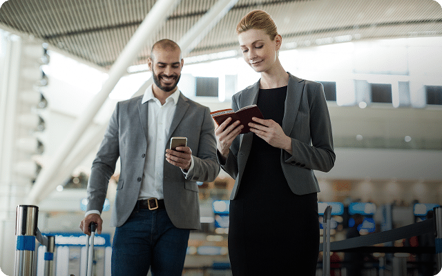 Two people in business attire at an airport terminal, one holding a passport and the other looking at a phone.